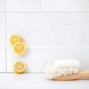 Close-up of shiny bathroom tiles with bright grout, lemon slices, baking soda, and a soft brush in a bright, airy bathroom.